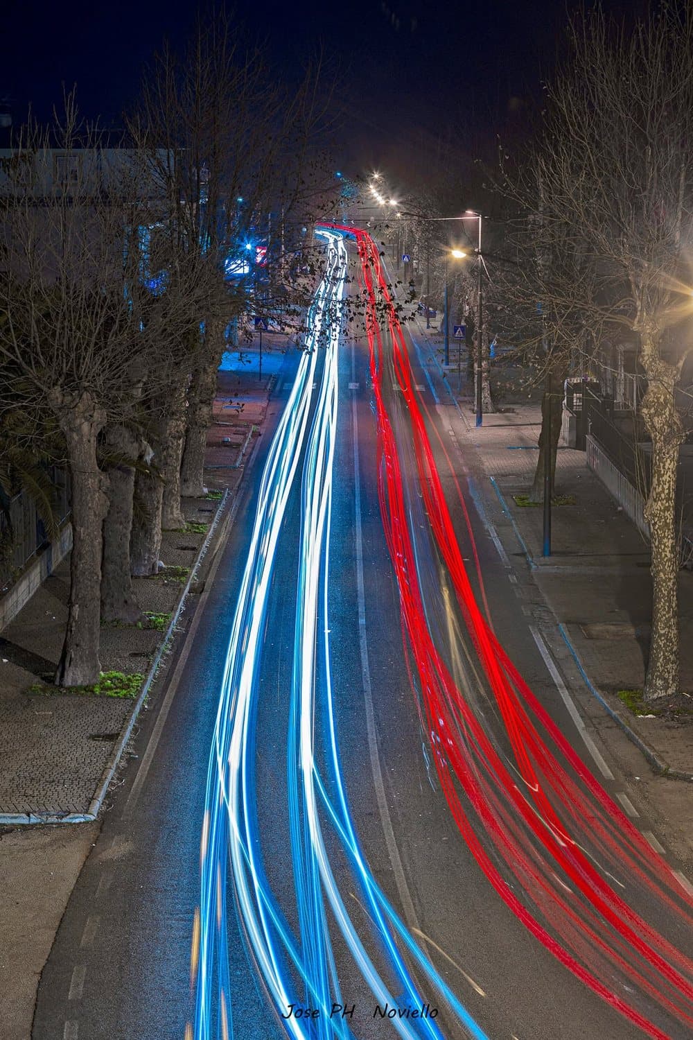 Strada di notte vista dall’alto con lunghe scie luminose bianche, blu e rosse lasciate dalle auto in movimento